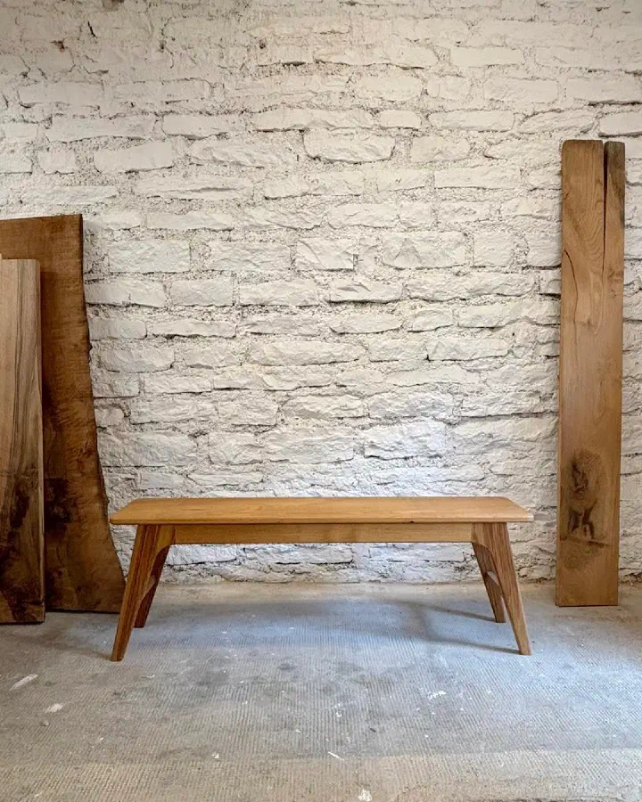 Banc en châtaignier massif à l'atelier, devant un mur de pierres blanches et des plateaux de bois brut