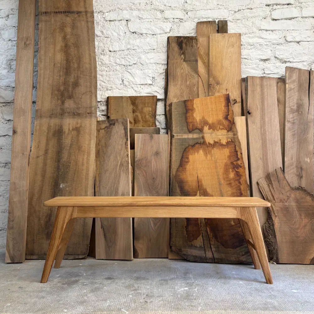 Banc en châtaignier massif vu de face, devant des planches de bois brut appuyées contre un mur de briques blanches
