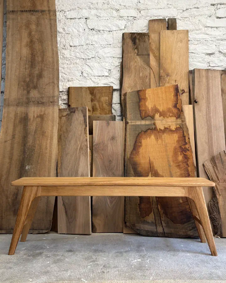 Banc en châtaignier massif vu de face, devant des planches de bois brut appuyées contre un mur de briques blanches