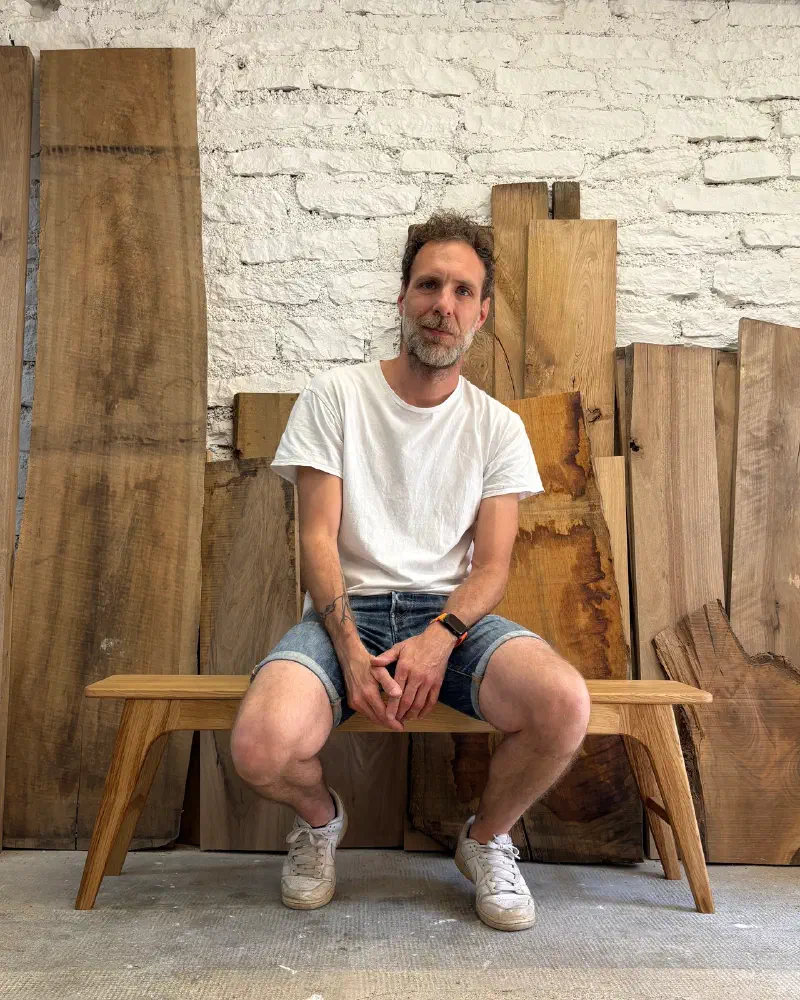 Benoit Guerriero assis sur un banc en chêne dans son atelier, devant des planches de bois brut