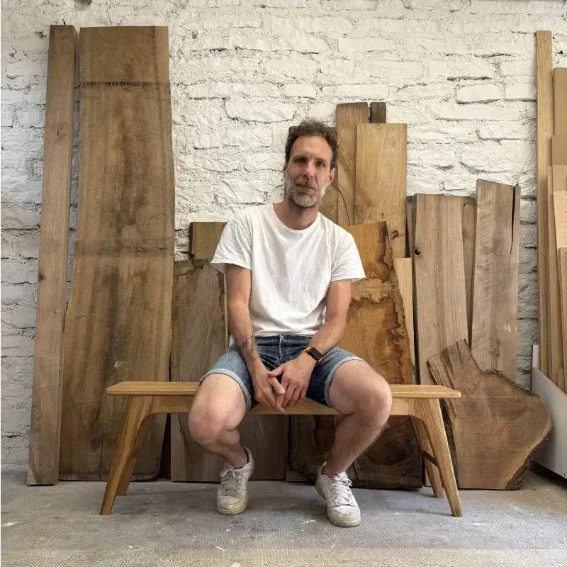 Benoit Guerriero assis sur un banc en chêne dans son atelier, devant des planches de bois brut