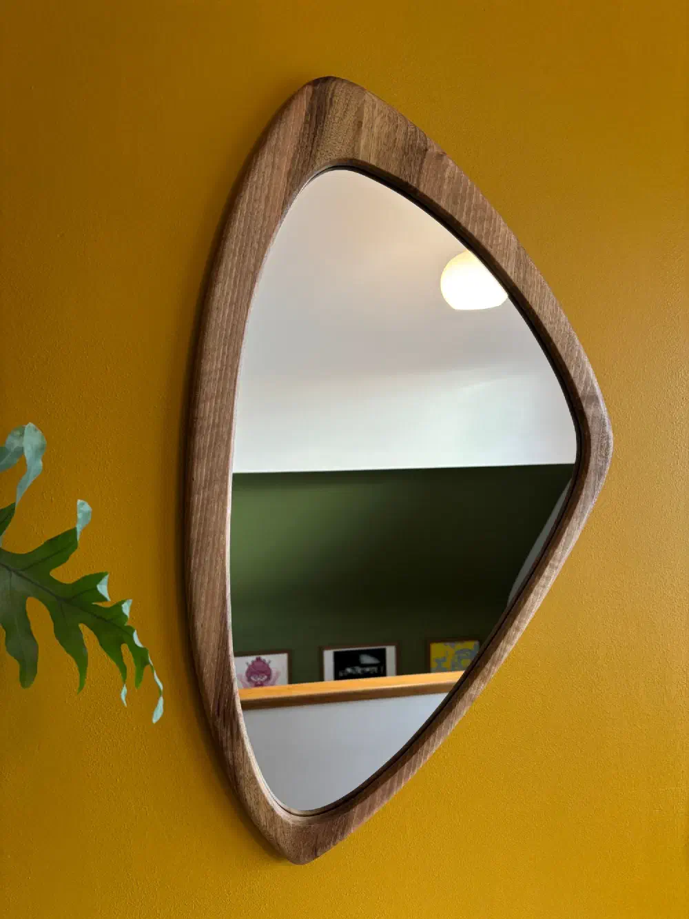Miroir en noyer massif à la forme asymétrique, accroché sur un mur jaune moutarde, à côté d'une feuille de philodendron