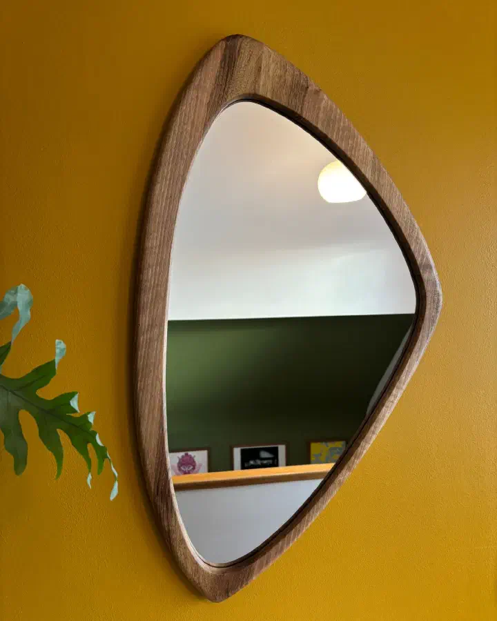 Miroir en noyer massif à la forme asymétrique, accroché sur un mur jaune moutarde, à côté d'une feuille de philodendron