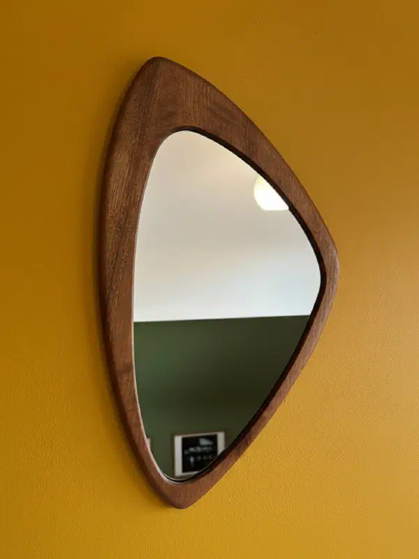 Petit miroir à la forme asymétrique, cadre en noyer massif, sur un mur jaune