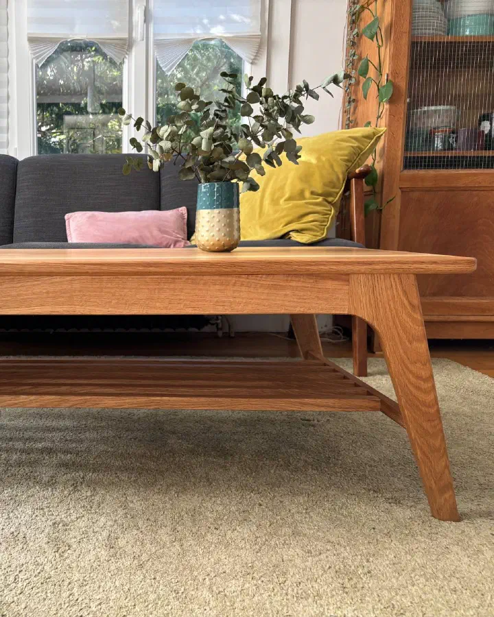 Table basse en chêne vue de face devant un canapé : ceinture, pieds fuselés et inclinés, tablette à claire-voie