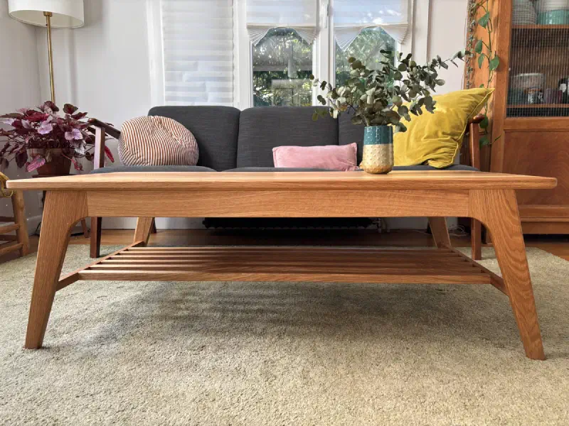 Table basse en chêne vue de face devant un canapé : ceinture, pieds fuselés et inclinés, tablette à claire-voie