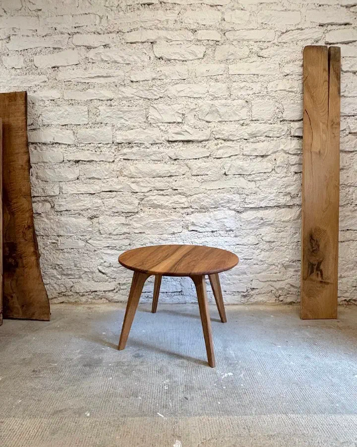 Petite table basse ronde en noyer massif à l'atelier, devant un mur de pierres blanches et des plateaux de bois brut