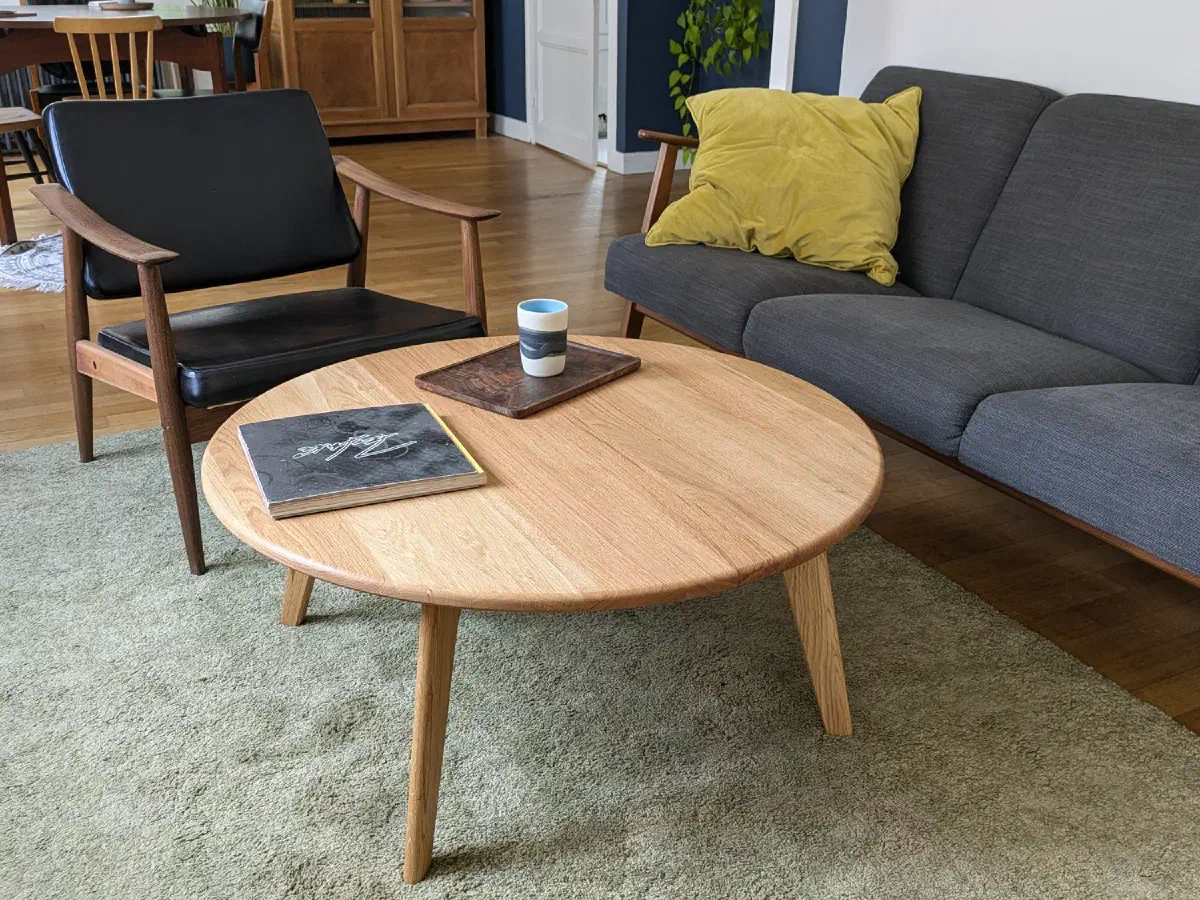 Grande table basse ronde en chêne dans un salon, entre un canapé gris et un fauteuil en cuir noir