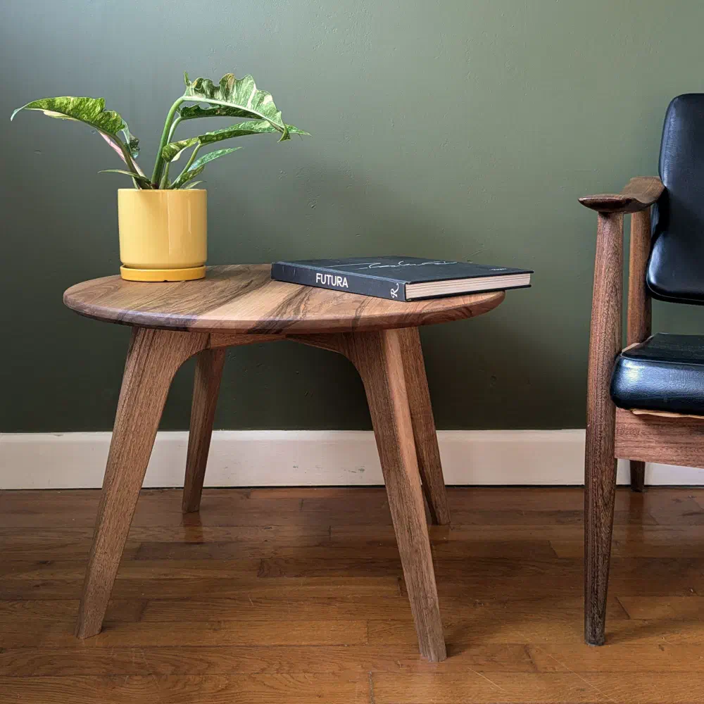 Petite table basse ronde en noyer massif à côté d'un fauteuil en cuir noir, devant un mur vert olive, une plante dans un pot jaune et un livre posés dessus