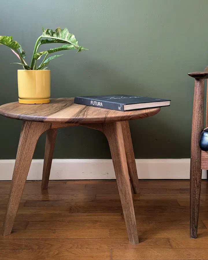 Petite table basse ronde en noyer massif à côté d'un fauteuil en cuir noir, devant un mur vert olive, une plante dans un pot jaune et un livre posés dessus